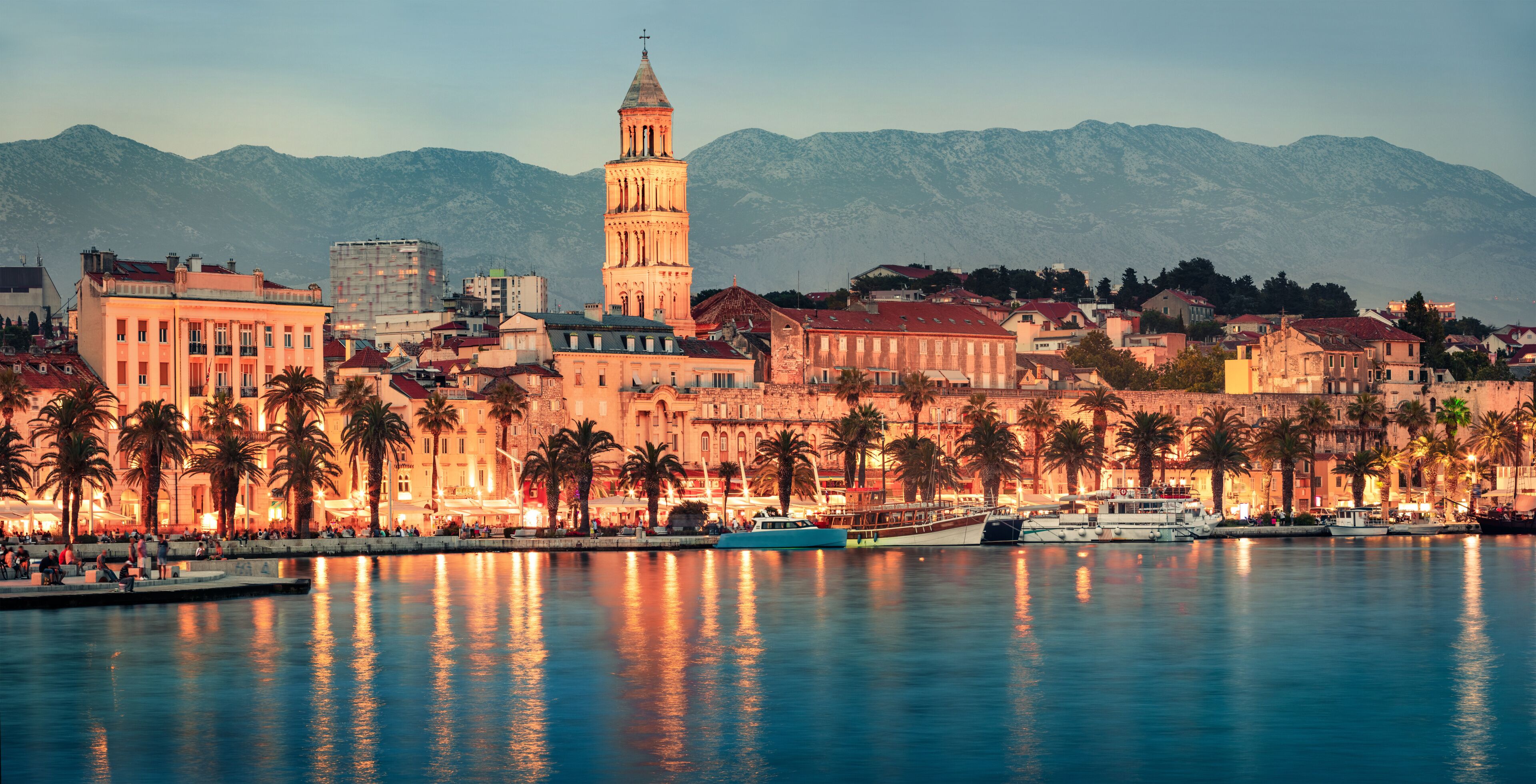 A coastal cityscape with a prominent tower, palm-lined waterfront, and mountains in the background.