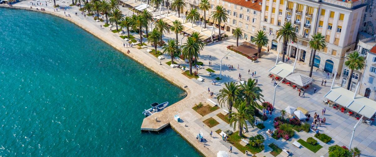 A coastal cityscape with a waterfront promenade lined with palm trees and historic buildings.
