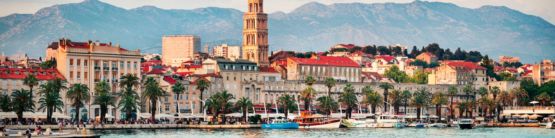 A coastal city with a prominent tower, a calm harbor with boats, and a backdrop of mountains.