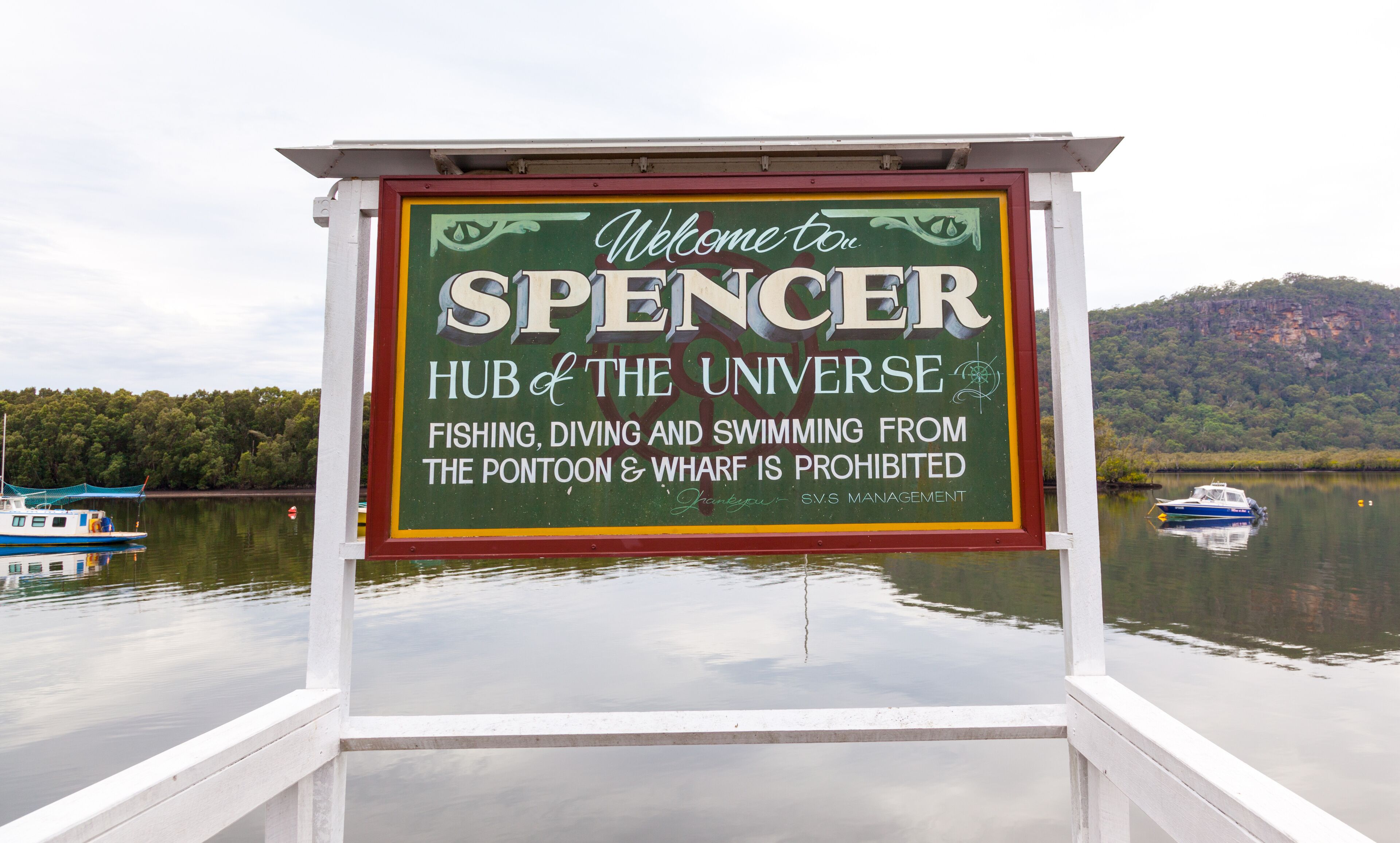 A sign welcoming visitors to Spencer, Hub of the Universe, with a lake and boats in the background.