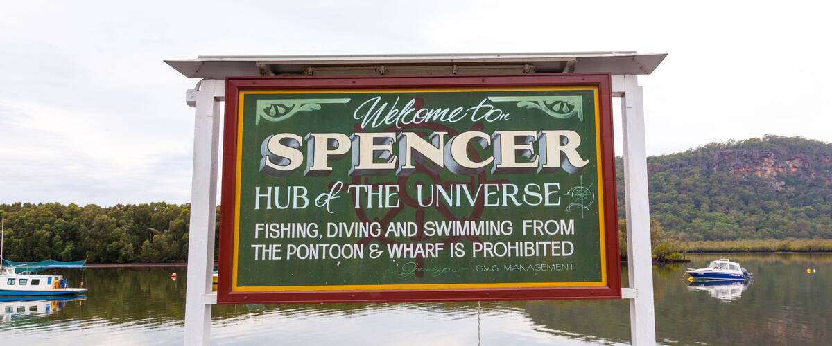 A sign welcoming visitors to Spencer, Hub of the Universe, with a lake and boats in the background.