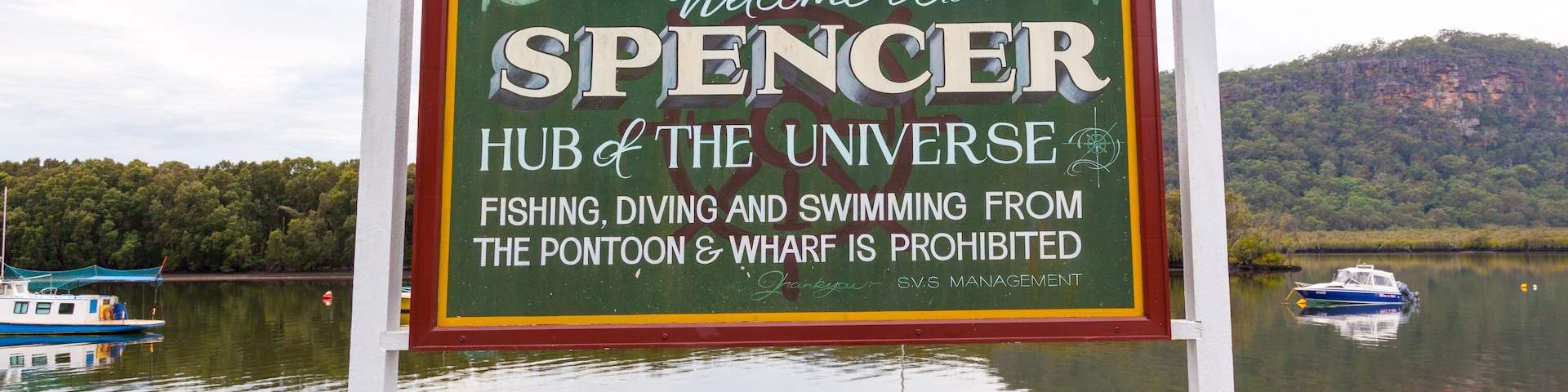 A sign welcoming visitors to Spencer, Hub of the Universe, with a lake and boats in the background.