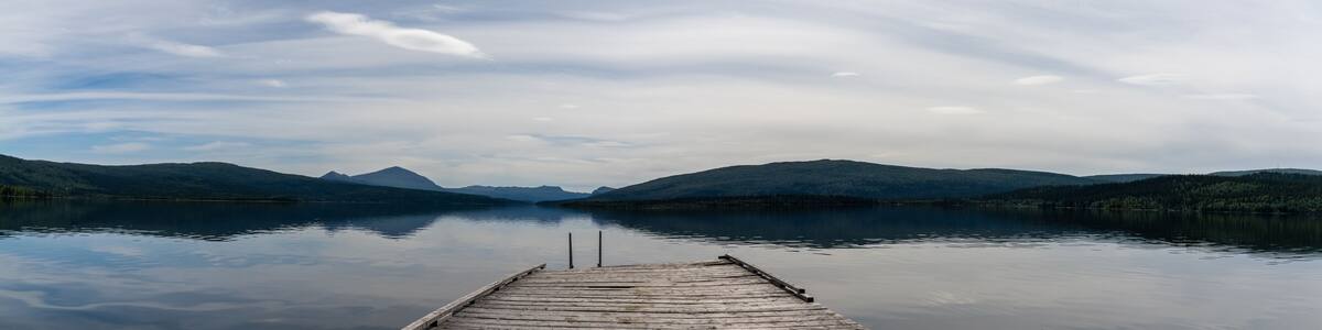 panorama of a calm lake with reflections of mountains and sky and a wooden dock in the foreground