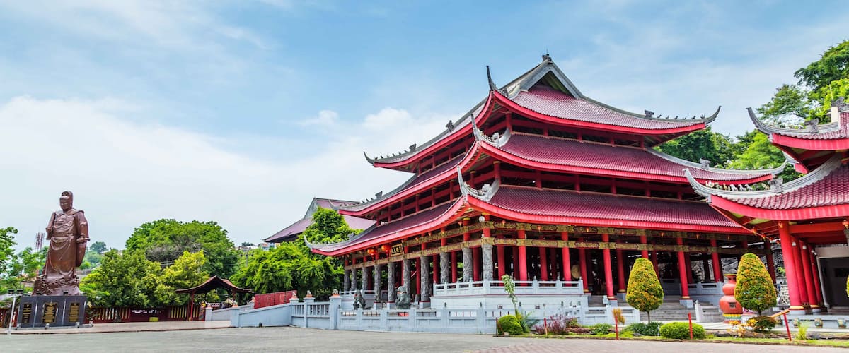 A traditional multi-tiered pagoda with red and gold accents, surrounded by lush greenery and a statue.