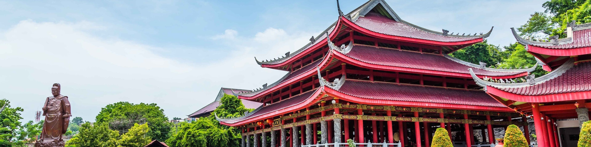 A traditional multi-tiered pagoda with red and gold accents, surrounded by lush greenery and a statue.