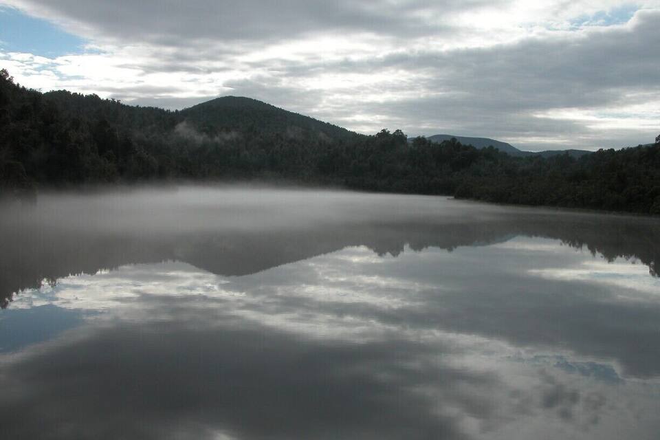 A calm lake reflecting clouds and a forested hill.