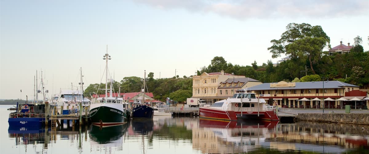 A harbor with several boats docked, a large building with a red roof, and a hotel in the background.