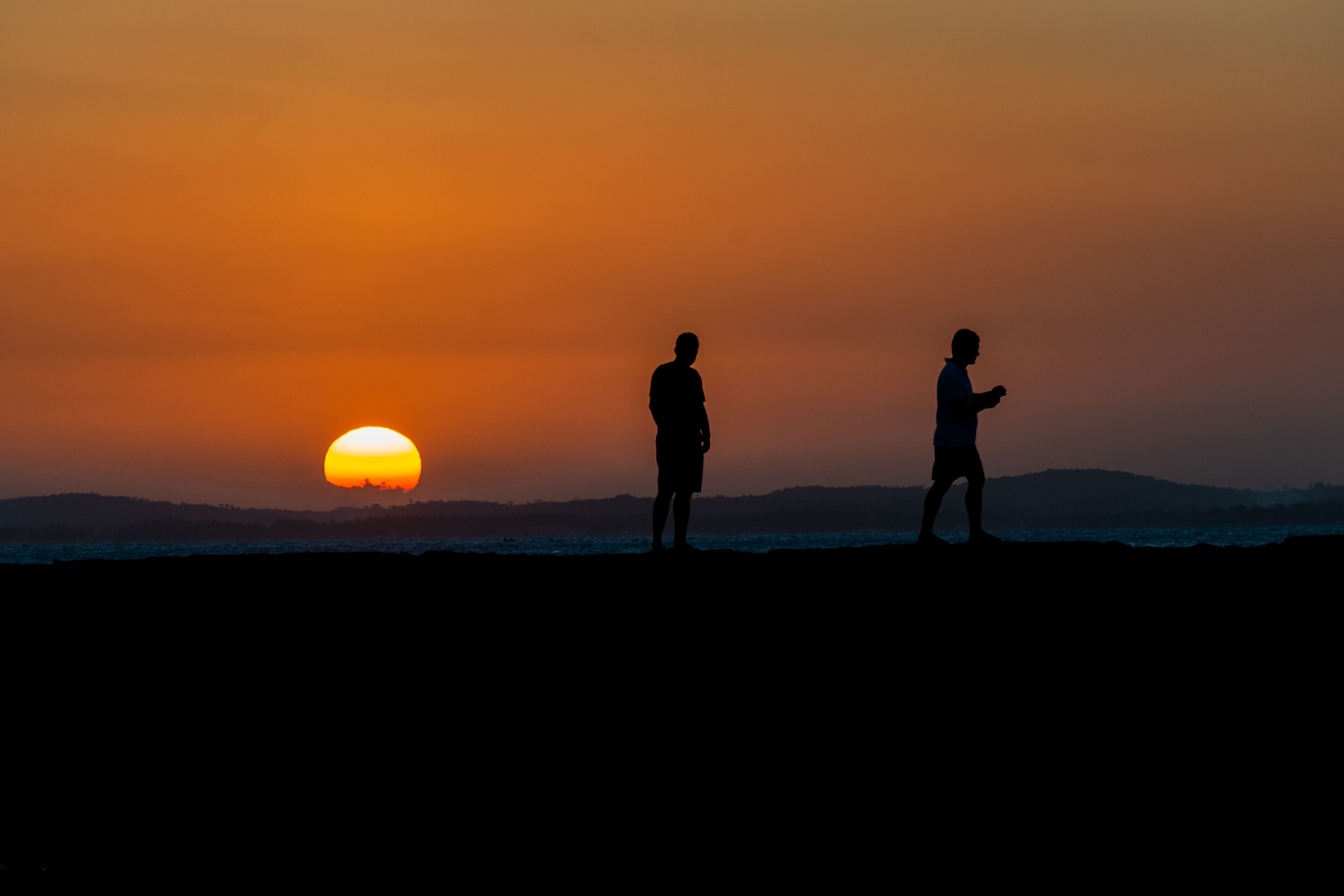 Dos siluetas de personas de pie en una playa con un atardecer de fondo.