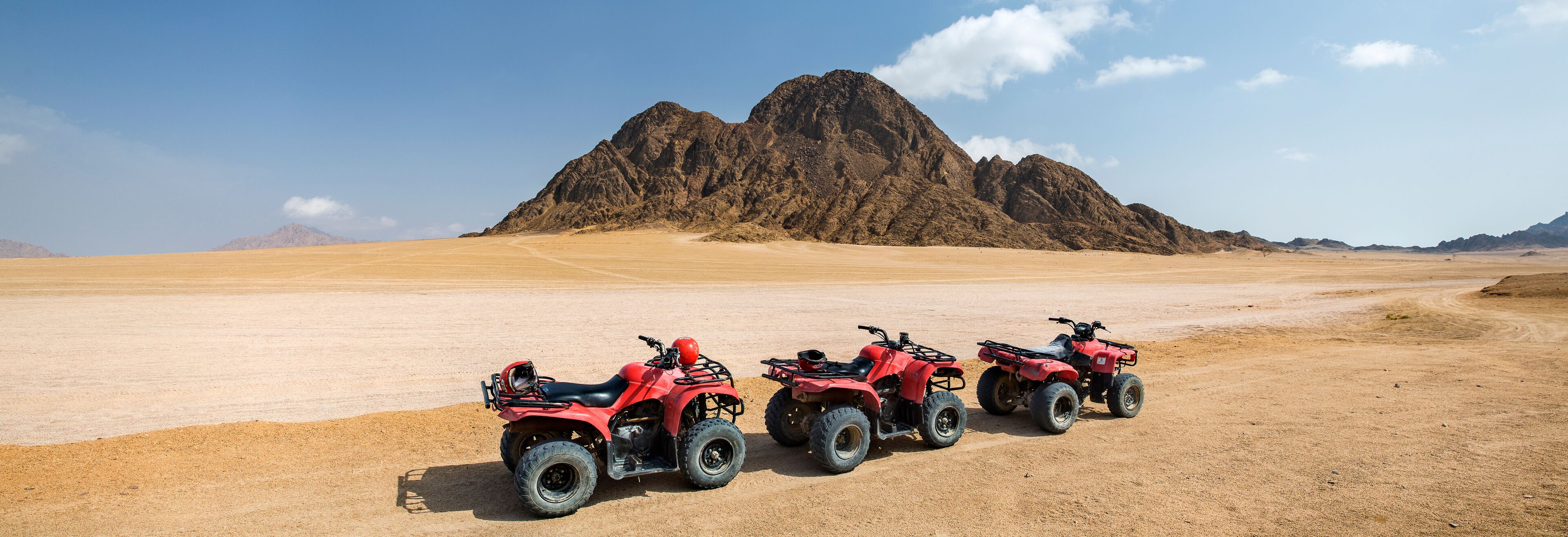 Three red ATVs parked on a desert landscape with a large mountain in the background.