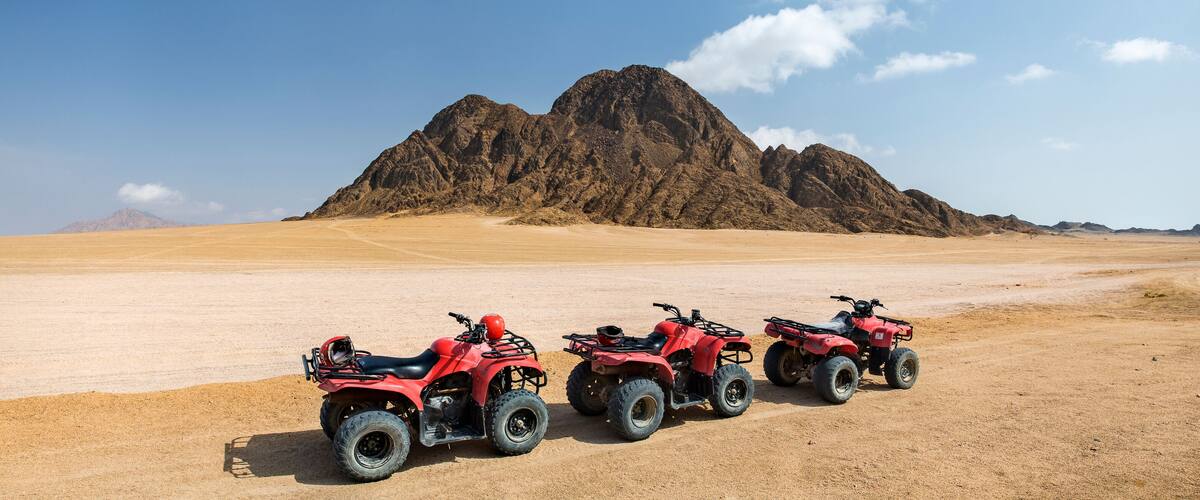 Three red ATVs parked on a desert landscape with a large mountain in the background.