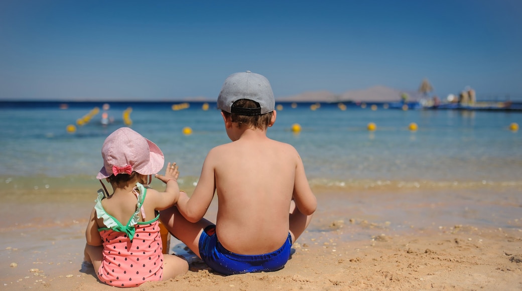 back view photo of a little girl in swimsuit and pink hat sitting on the beach with her elder brother with a cap on his head