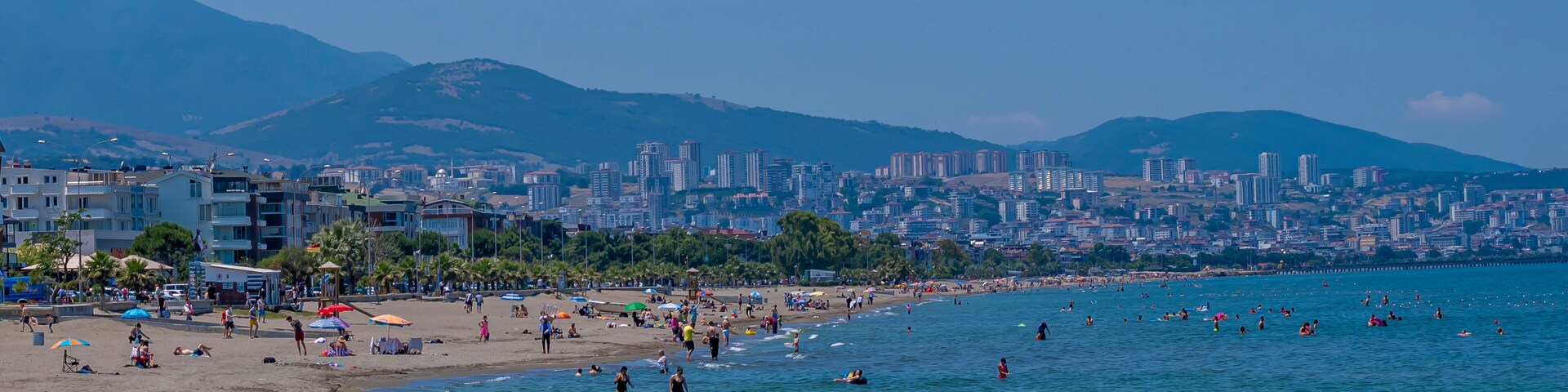 Uma praia com pessoas, paisagem urbana e montanhas ao fundo.