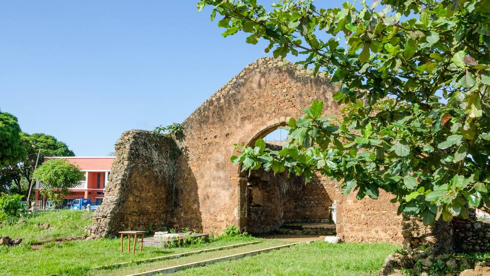 An ancient stone archway surrounded by greenery, with a railway track in the foreground and a building in the background.