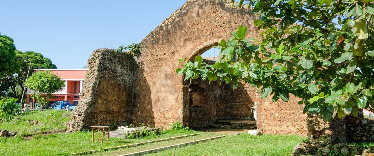 An ancient stone archway surrounded by greenery, with a railway track in the foreground and a building in the background.