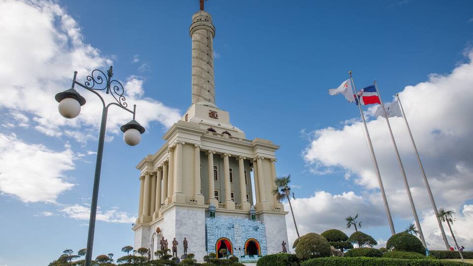 Un monumento con una alta columna y una cruz en la parte superior, rodeado de estatuas y banderas.