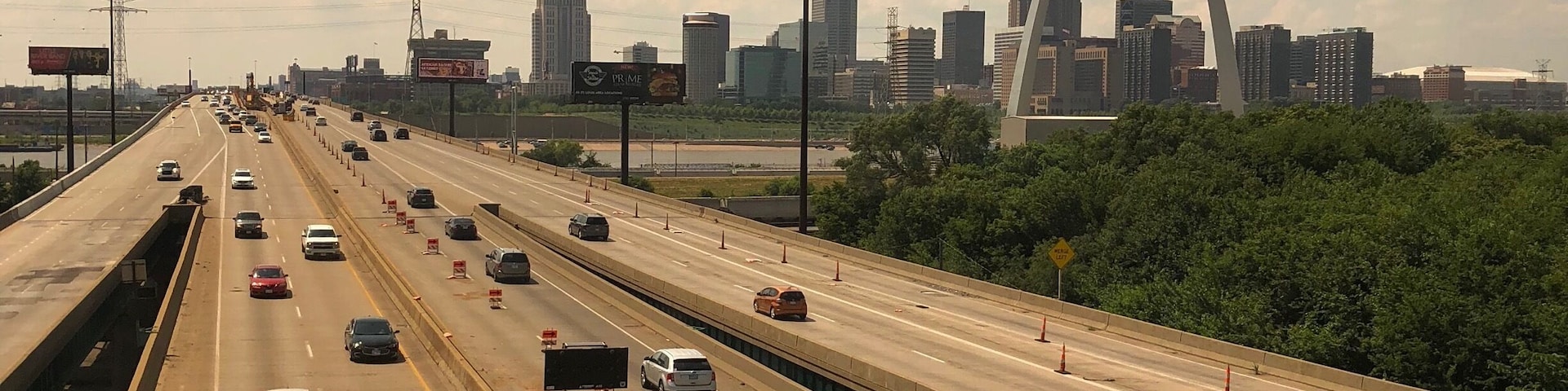 A multi-lane highway with vehicles, a city skyline in the background, and a distinctive arch bridge.