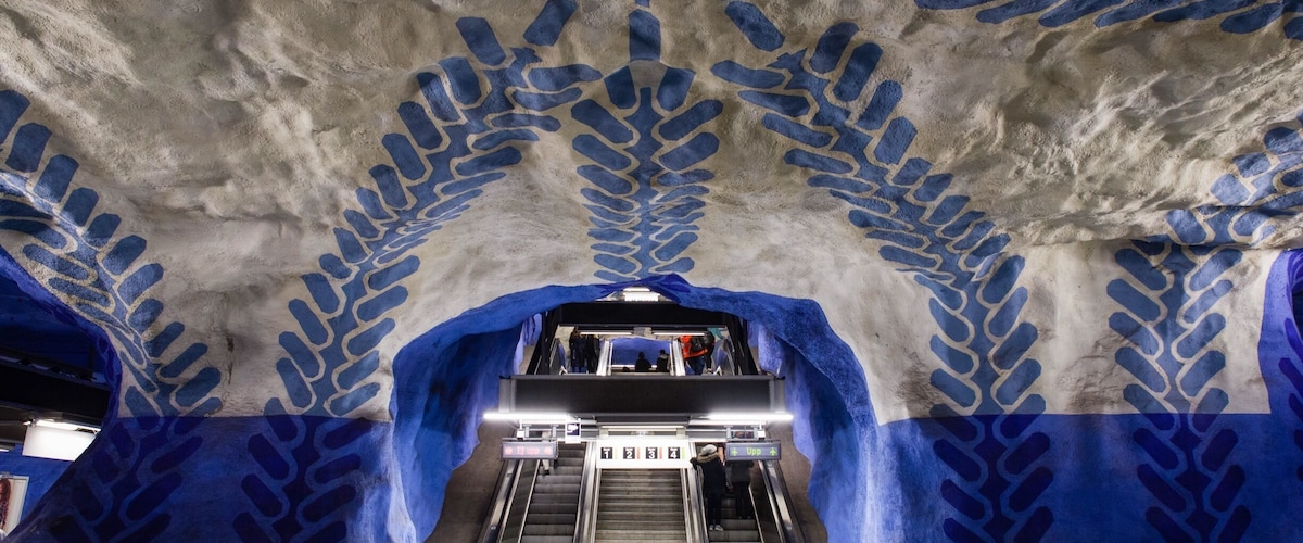 En tunnelbanestation med blå mönster på väggarna och taket, och rulltrappor som leder ner till perrongen.