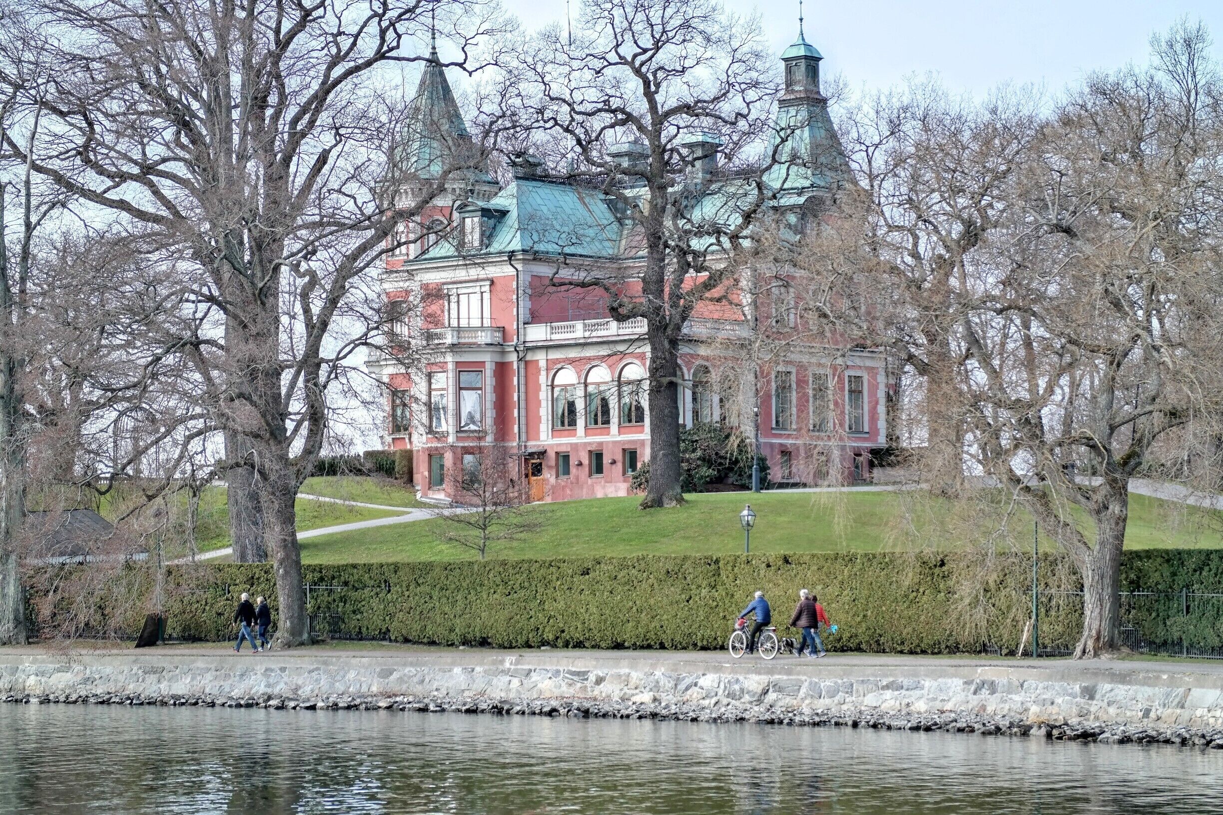 A historic building with red brick and green roof, surrounded by trees and a body of water.