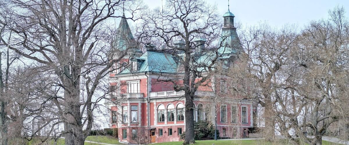 A historic building with red brick and green roof, surrounded by trees and a body of water.
