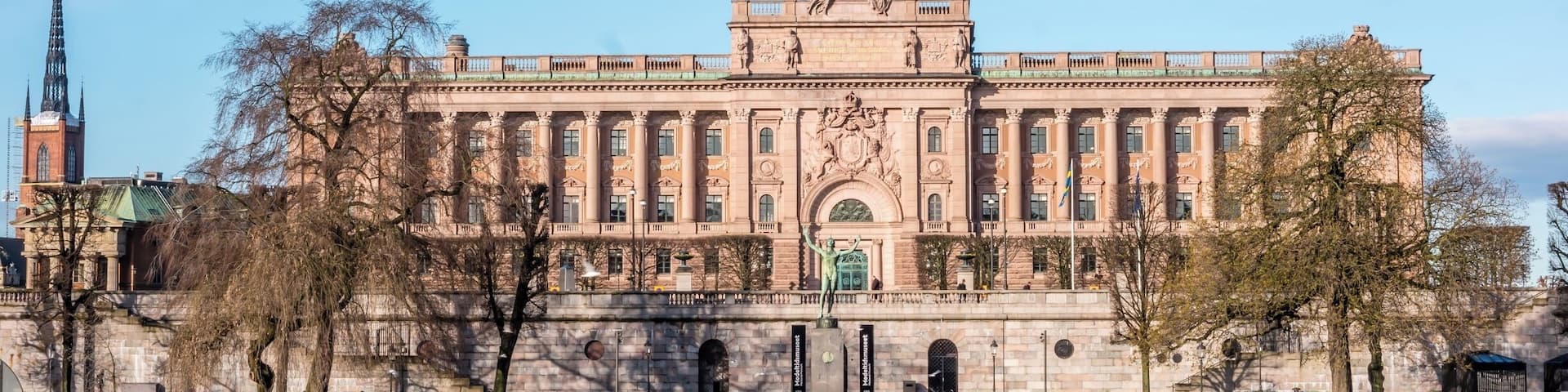 A large, historic building with a central statue and a river in the foreground.
