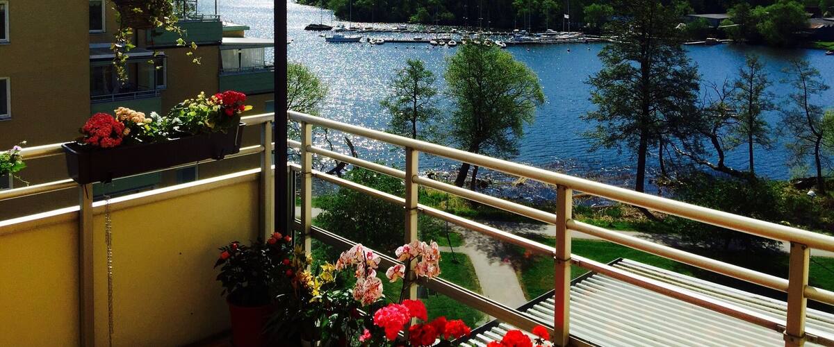 A balcony with potted flowers overlooking a river with boats and a forested hillside.