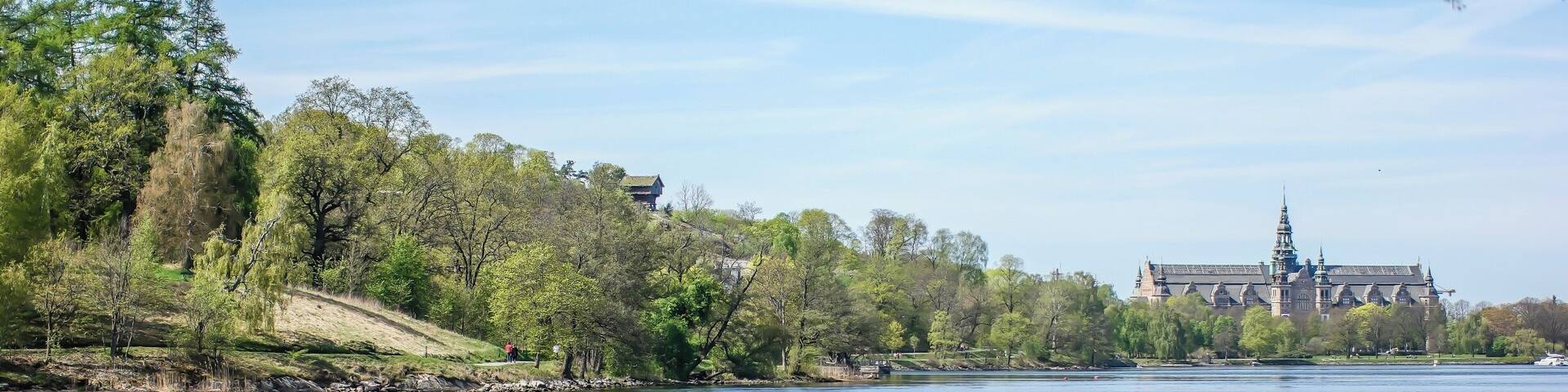A calm river with a boat carrying three people, surrounded by lush greenery and a historic building in the background.