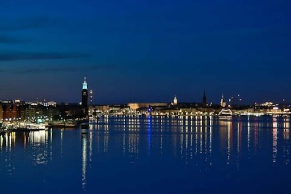 A nighttime cityscape with a prominent clock tower reflecting on calm water.