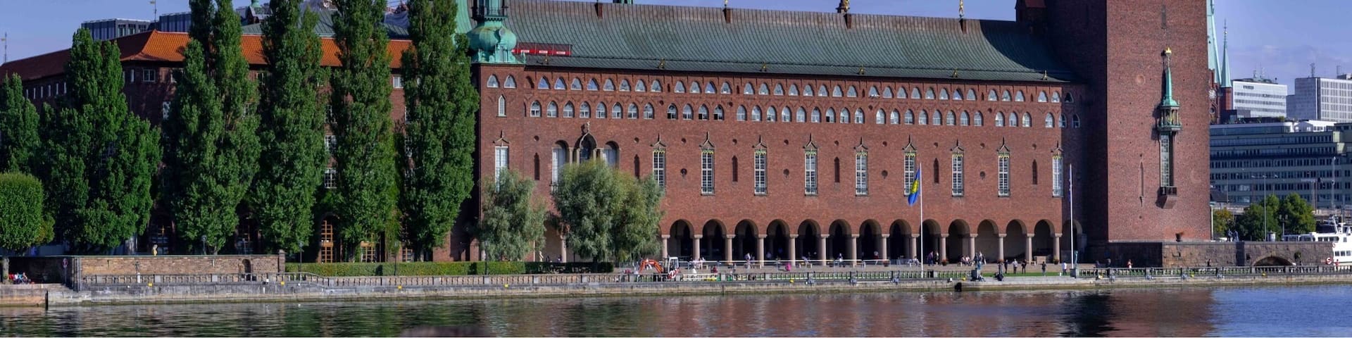 A large brick building with a tall tower and a green dome, located near a body of water.
