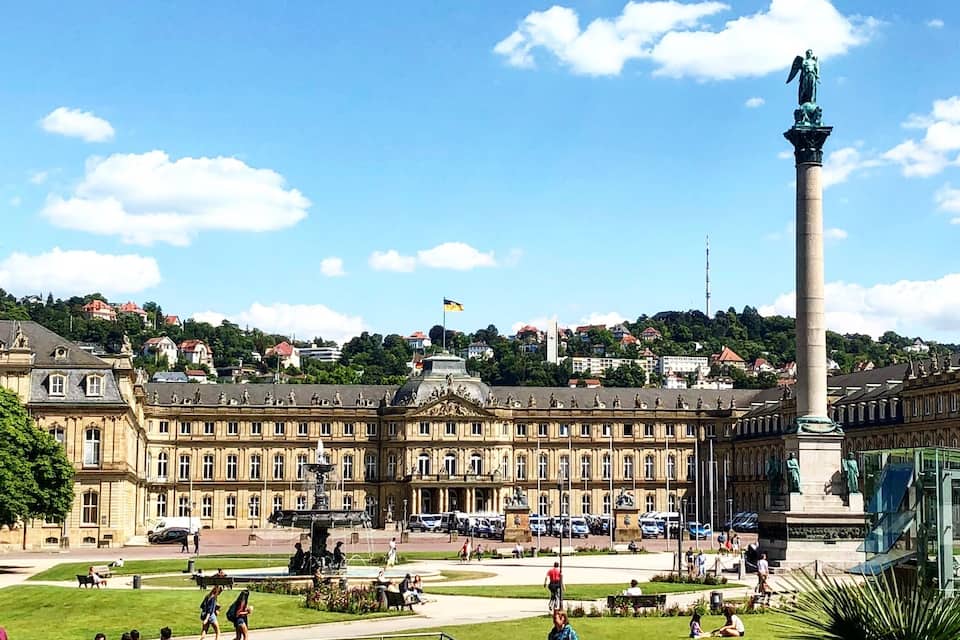 Un edifici gran i antic amb una font a la façana, envoltat d'un parc on hi passegen gent.