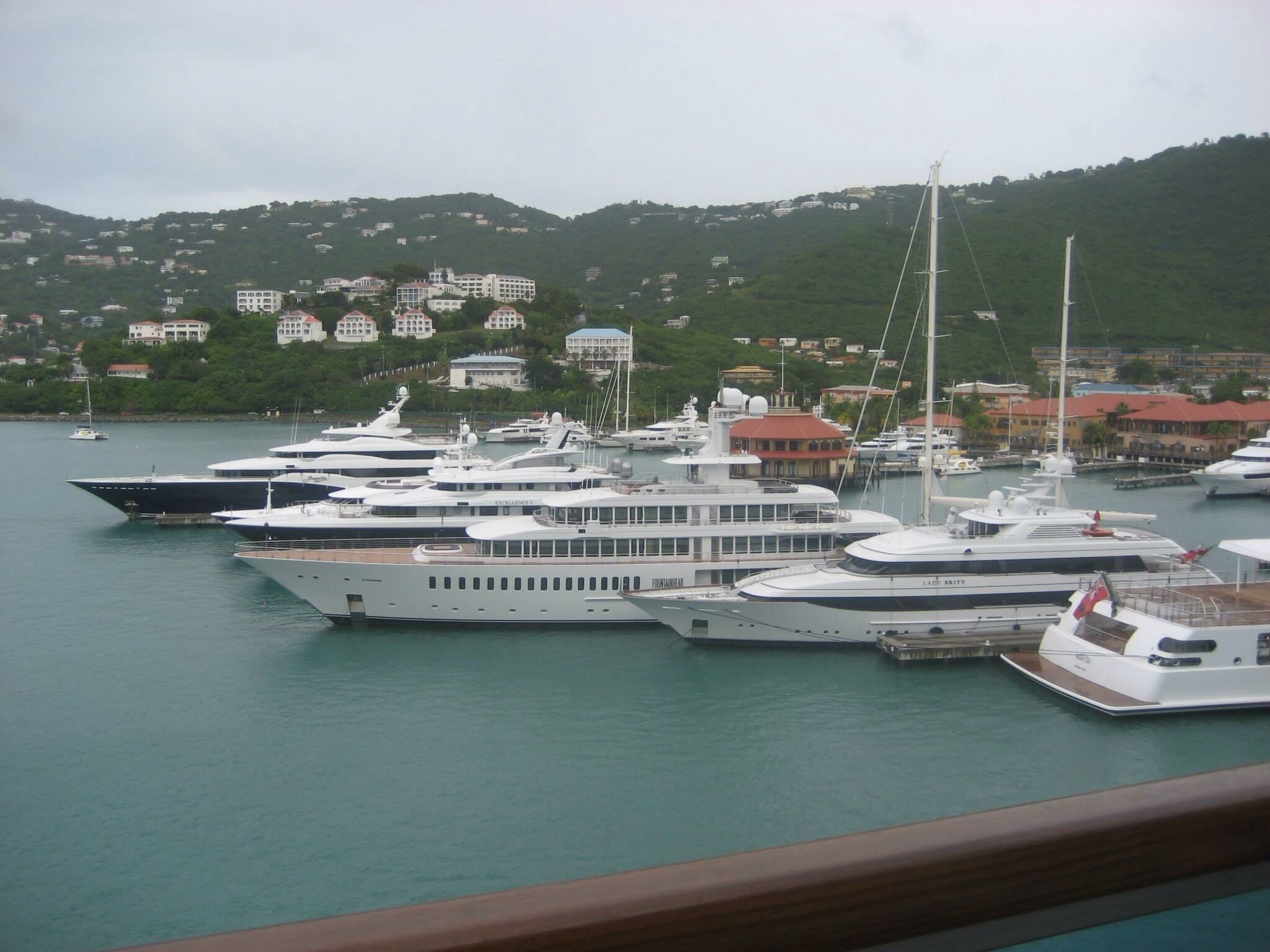A marina with multiple yachts docked, surrounded by a hilly landscape with buildings in the background.