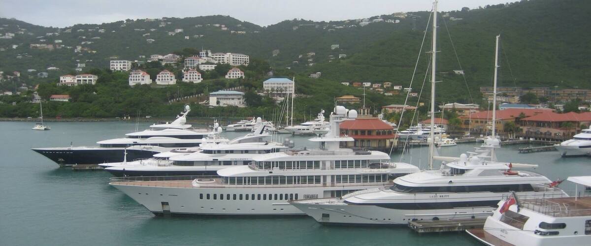 A marina with multiple yachts docked, surrounded by a hilly landscape with buildings in the background.
