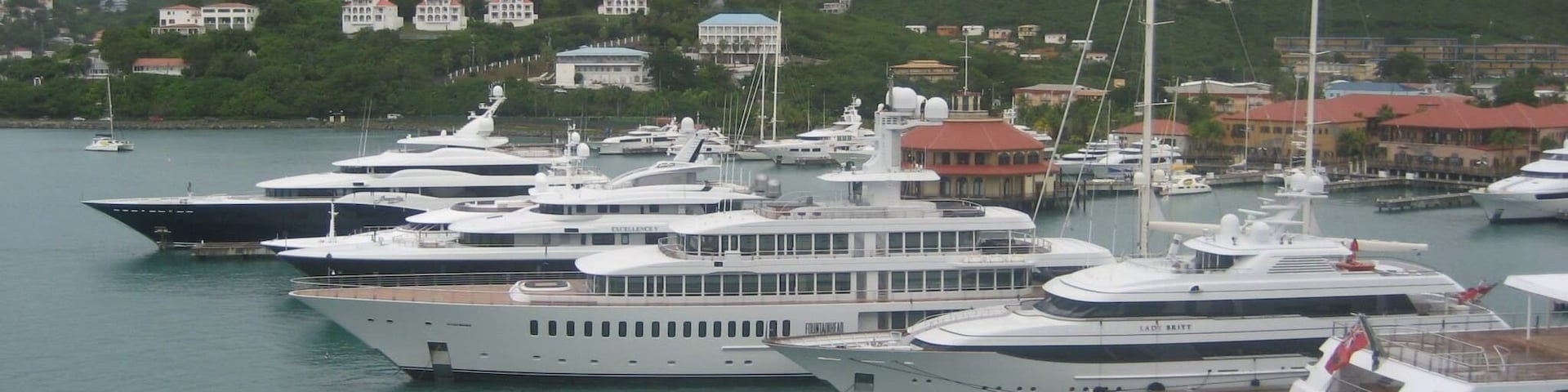 A marina with multiple yachts docked, surrounded by a hilly landscape with buildings in the background.