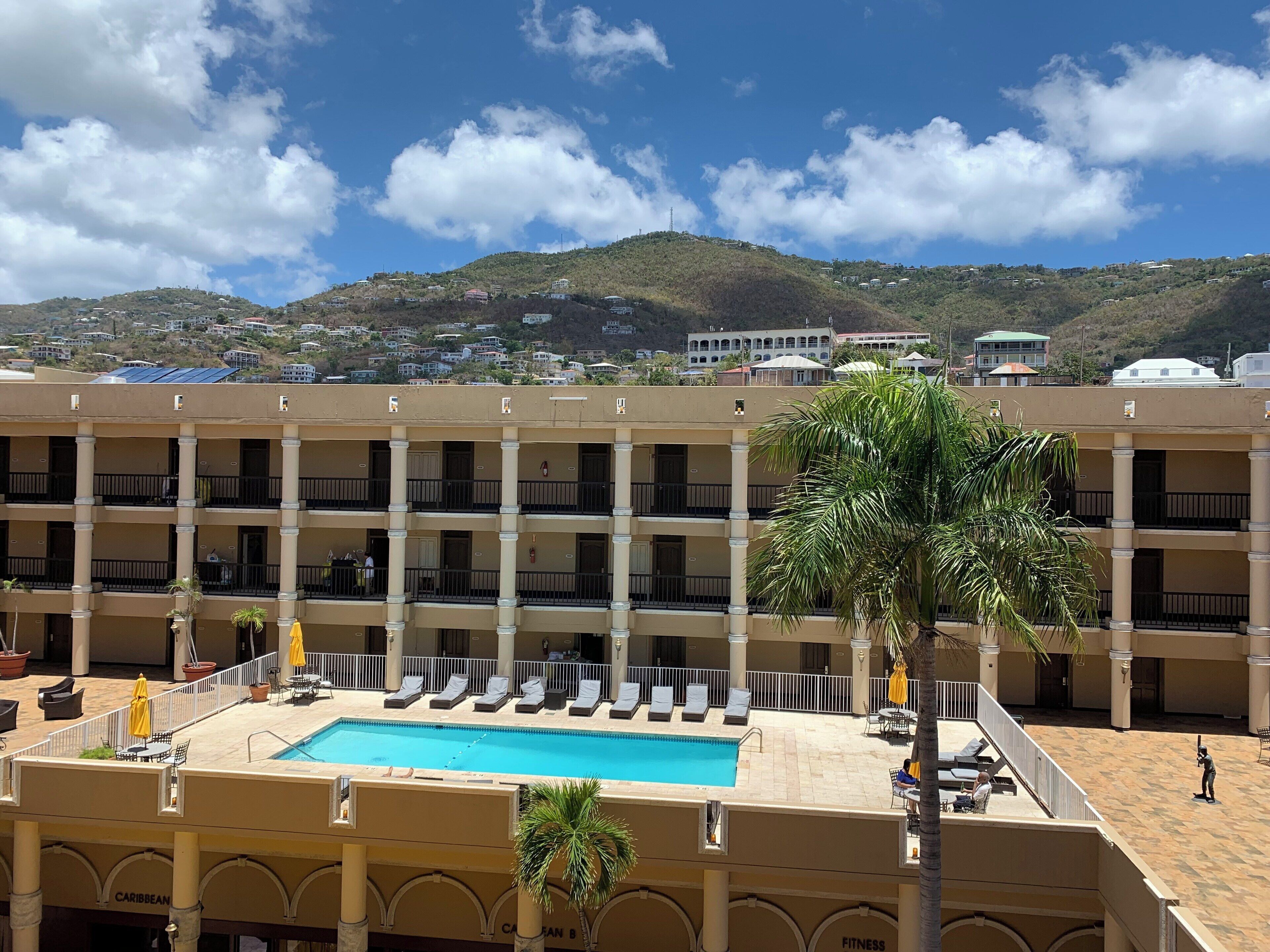 A hotel with a swimming pool, palm trees, and a view of a hilly town in the background.