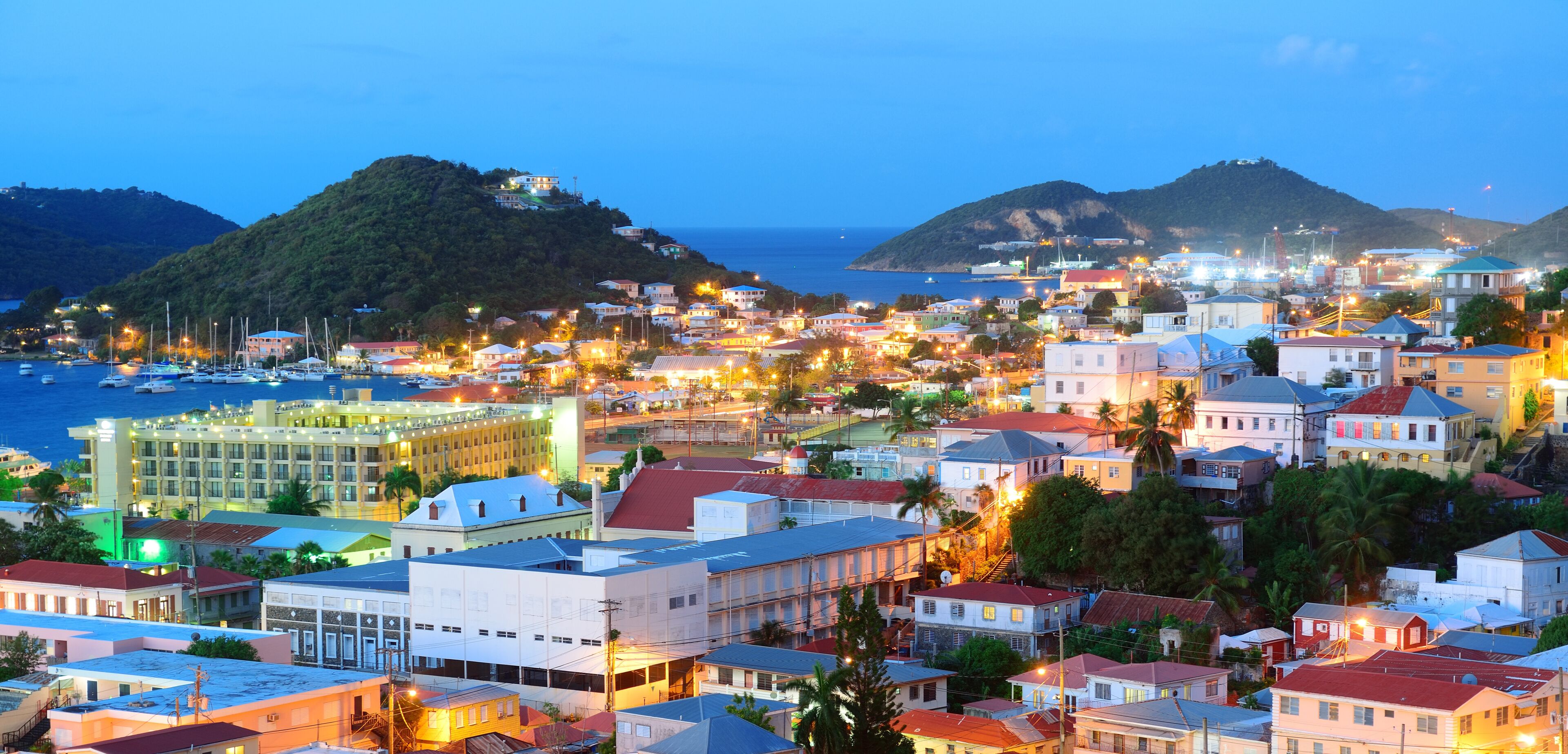 Uma vila costeira ao entardecer, com edifícios, um porto de iates e colinas ao fundo.
