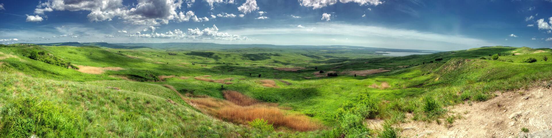 Rolling green hills, a clear blue sky with scattered clouds, and a distant body of water.