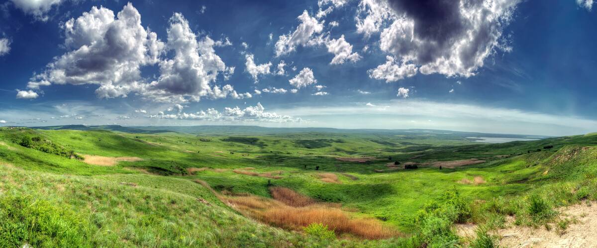 Rolling green hills, a clear blue sky with scattered clouds, and a distant body of water.