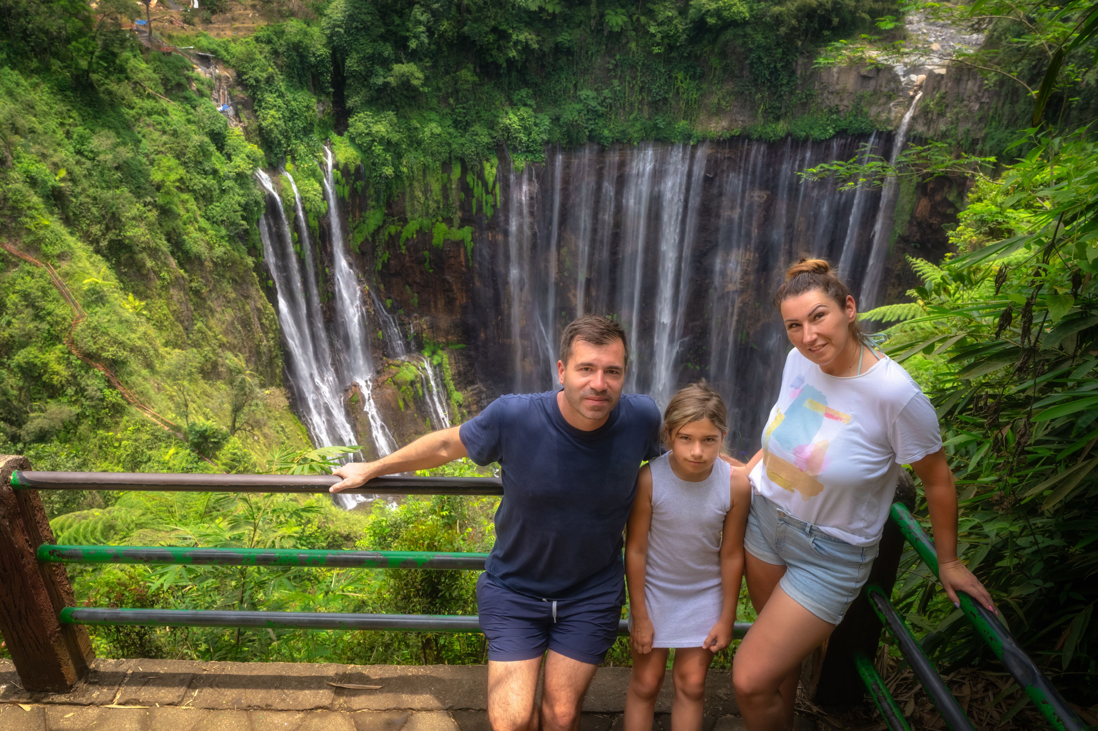 Uma cascata, vegetação exuberante e um corrimão.