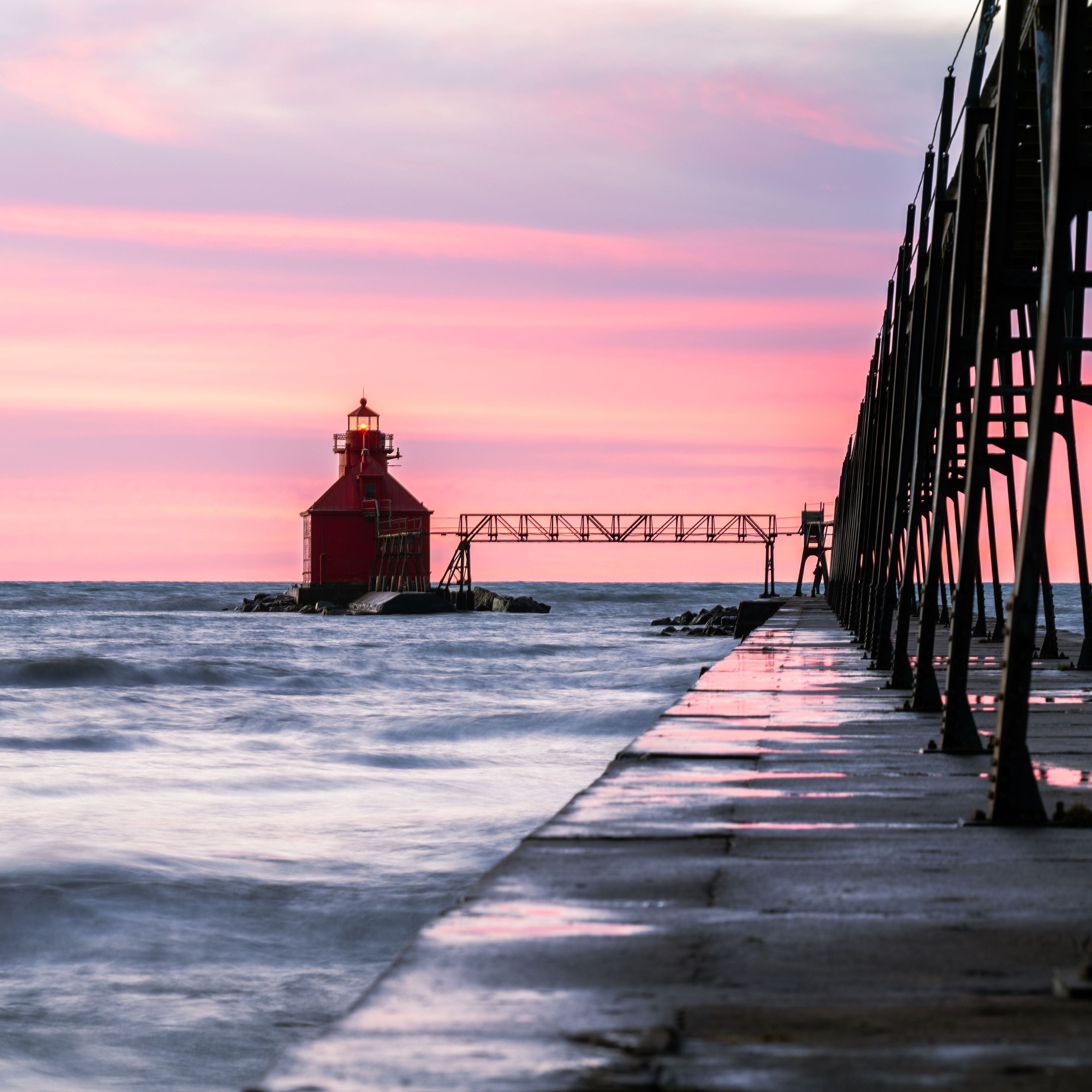 A red lighthouse at sunset, a pier extending into the water, and a calm sea.