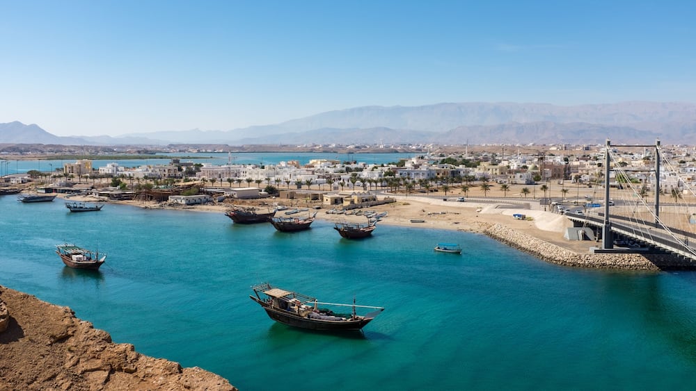 A coastal town with boats docked along the waterfront, a bridge, and mountains in the background.