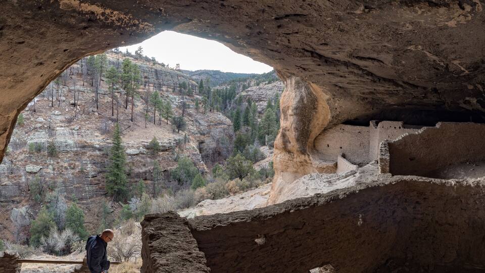 A cave with a natural archway, a person standing inside, and a view of a forested mountain outside.