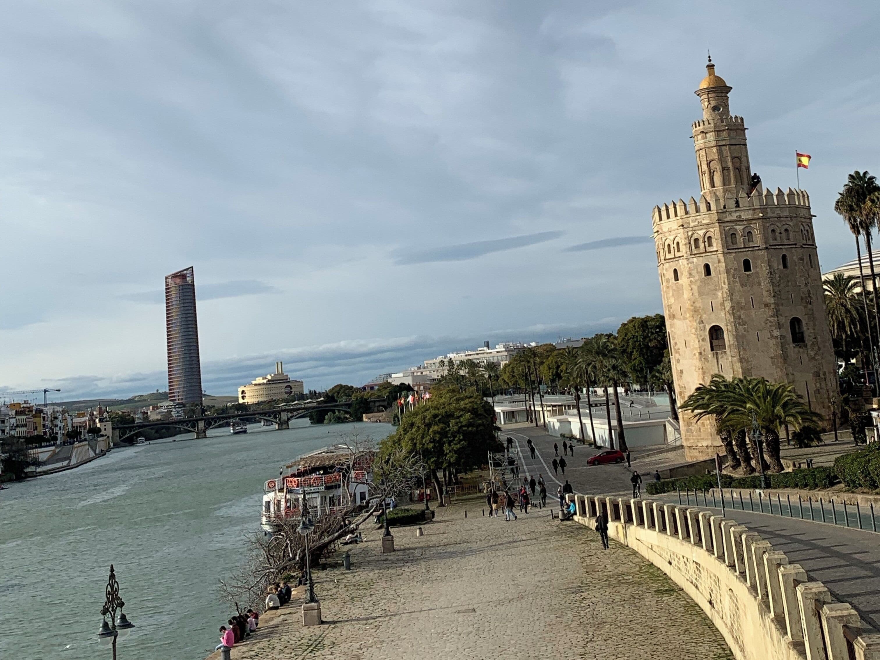 Un paseo ribereño con una torre histórica, un rascacielos moderno y un puente.