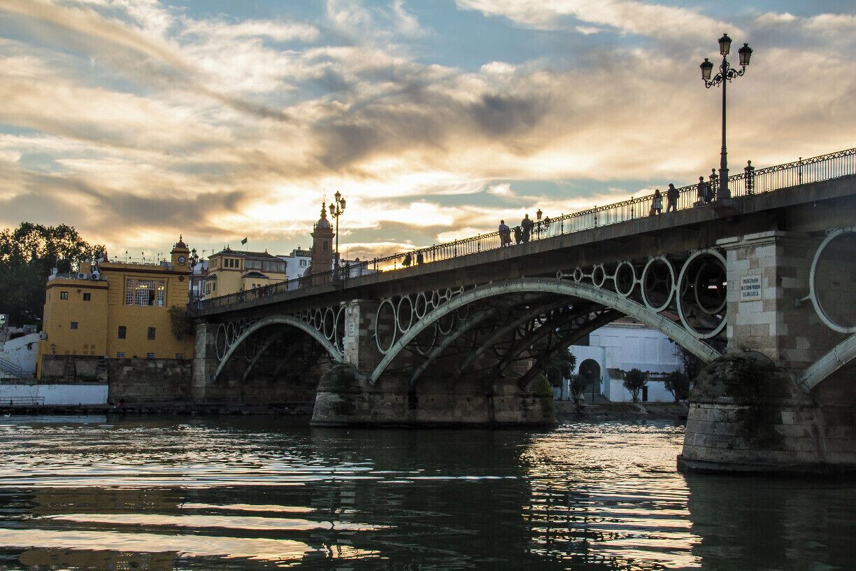 Un ponte di pietra storico con archi che sovrastano un fiume, circondato da edifici e un cielo nuvoloso.