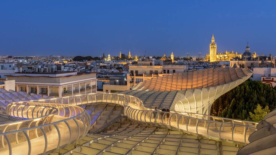 A modern stadium with a unique curved roof structure, surrounded by urban buildings and a prominent tower in the background.