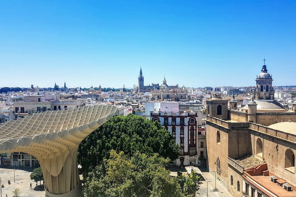 A cityscape with a prominent modern architectural structure in the foreground, historic buildings, and a clear blue sky.