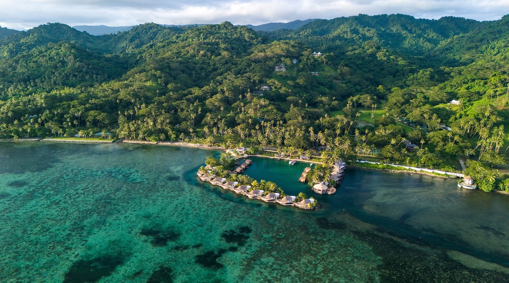 An aerial view of a tropical island with clear turquoise waters, a small island with overwater bungalows, and lush green mountains in the background.