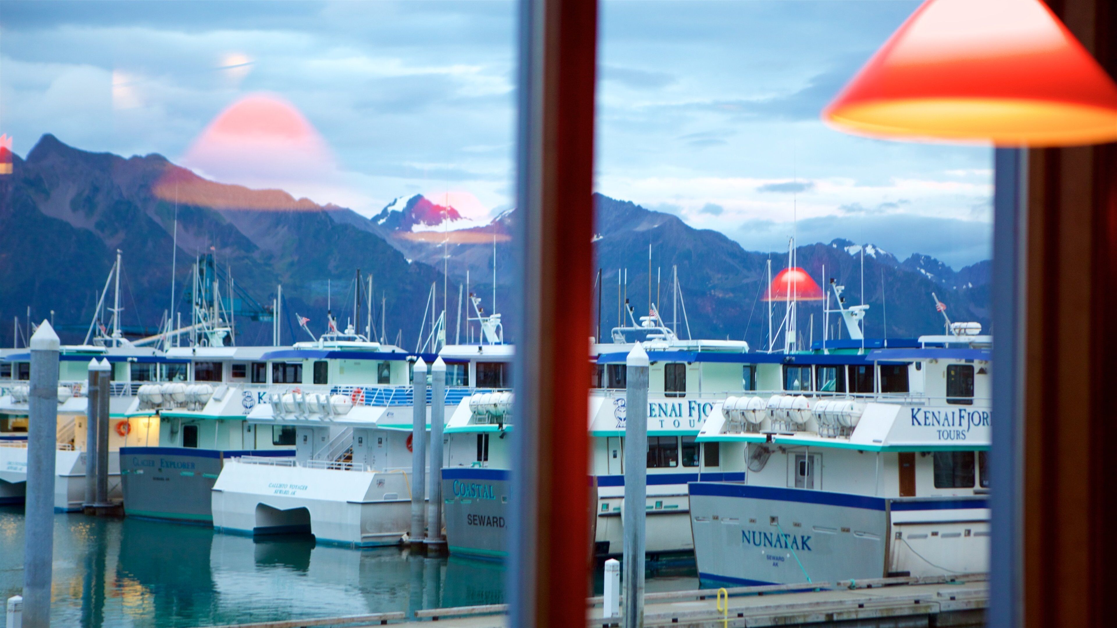 A marina with several boats docked, including 'Nunatak' and 'Kenai Fjords Tours'. Mountains are visible in the background.
