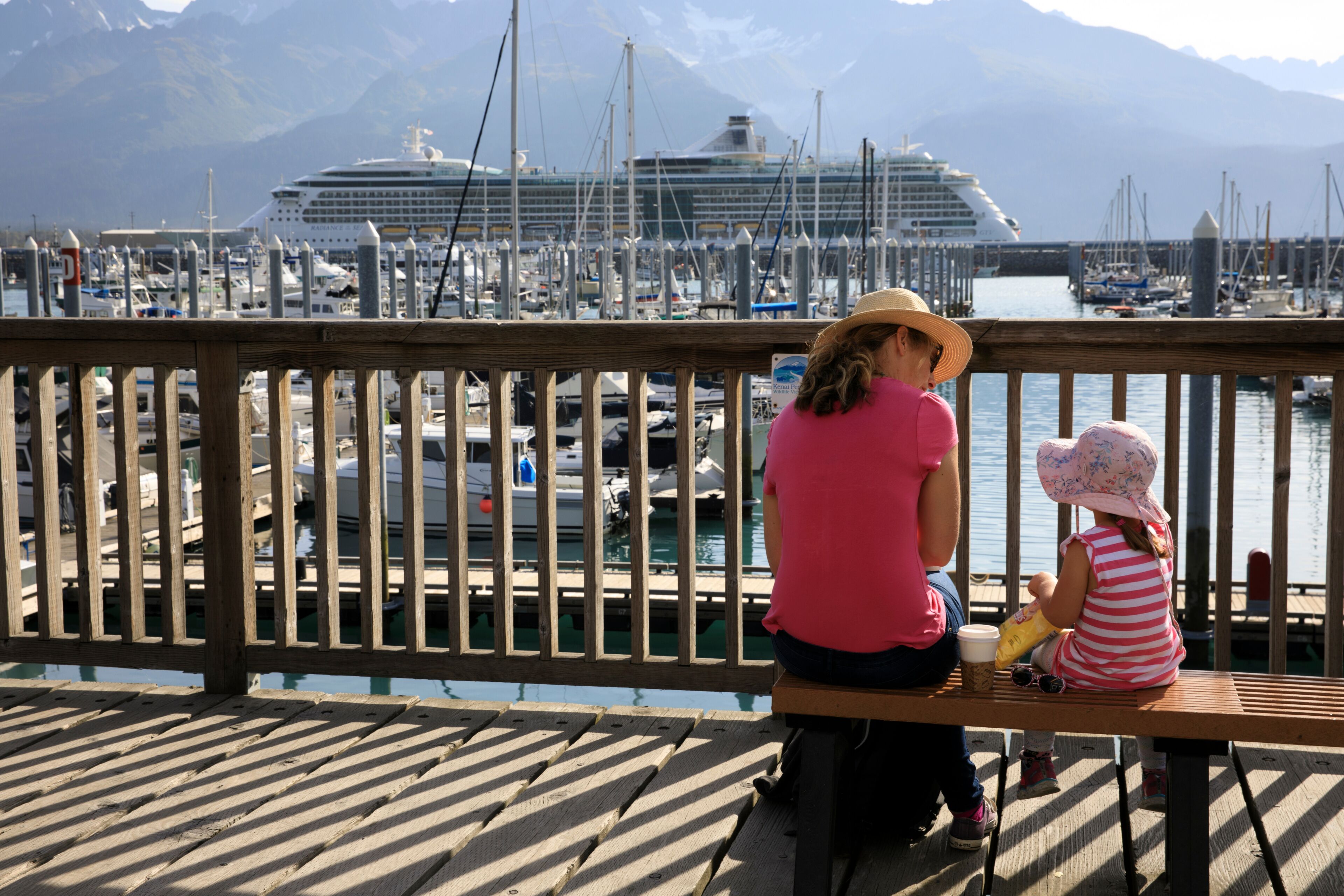Un muelle de madera con barandilla, una mujer y un niño sentados en un banco, un crucero y un puerto deportivo con barcos y montañas al fondo.