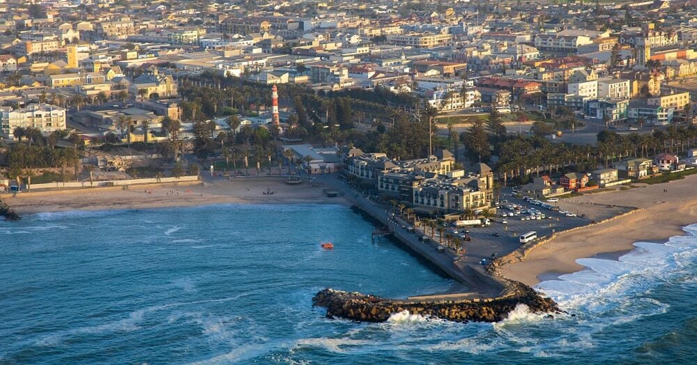 A coastal cityscape with a prominent lighthouse, a beach, and a rocky breakwater.