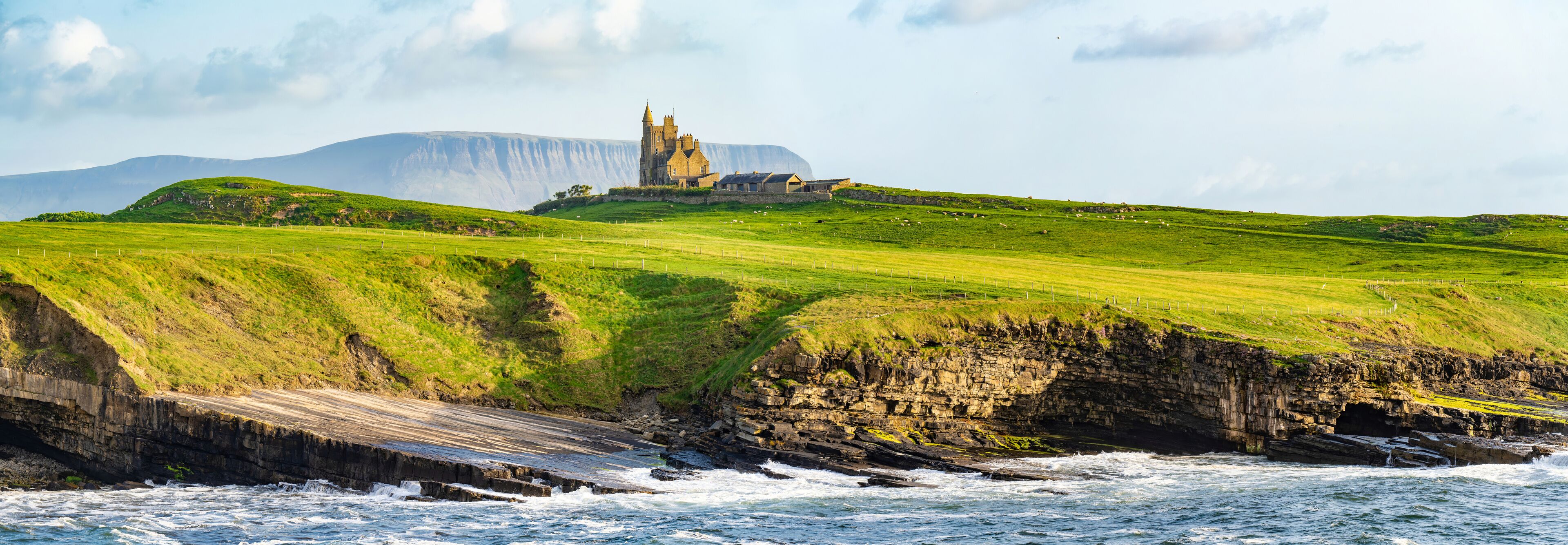 A coastal landscape with a castle on a grassy hill, waves crashing against the rocks, and a large mountain in the background.