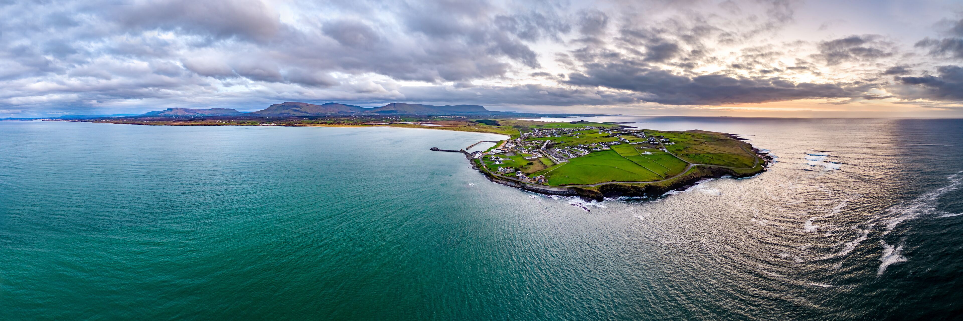 An aerial view of a coastal village with a small harbor, surrounded by mountains and a large body of water.